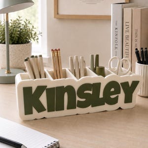 May include: A rectangular off-white desk organiser with five compartments, featuring the name Kinsley in large, raised, dark olive green letters on the front. The organiser holds various pens, pencils, and a pair of white scissors.