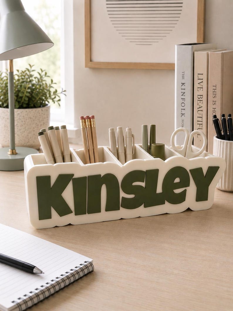 May include: A rectangular off-white desk organiser with five compartments, featuring the name Kinsley in large, raised, dark olive green letters on the front. The organiser holds various pens, pencils, and a pair of white scissors.