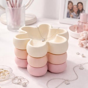 May include: A flower-shaped jewelry holder in cream, peach, and pink tones, with a silver necklace featuring a small, square pendant. Other silver necklaces and rings are also visible, along with makeup brushes and a framed photo.