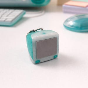 May include: A miniature teal and white computer keychain. The small, square-shaped keychain has a gray screen and teal accents. A silver chain is attached to the top. Other teal computer accessories are in the background.