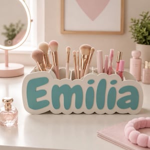 May include: A white desk organiser with a scalloped edge and the name Emilia in large teal letters on the front. The organiser contains various makeup brushes with light pink handles and several cosmetic tubes in shades of pink.