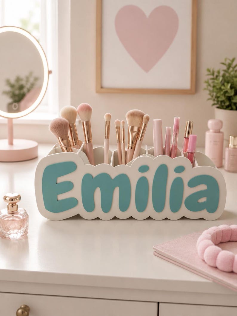 May include: A white desk organiser with a scalloped edge and the name Emilia in large teal letters on the front. The organiser contains various makeup brushes with light pink handles and several cosmetic tubes in shades of pink.