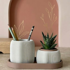 May include: A white ribbed desk organizer set with a matching tray. The set includes a pencil holder with pencils and a small potted succulent. The background features a pink arch and gold leaf design.