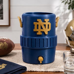 May include: A blue cylindrical beverage cooler featuring a gold Notre Dame ND logo on the front, two gold side handles, and a gold spigot at the base. The cooler has a ribbed texture around the middle section and a smooth finish on the top and bottom.