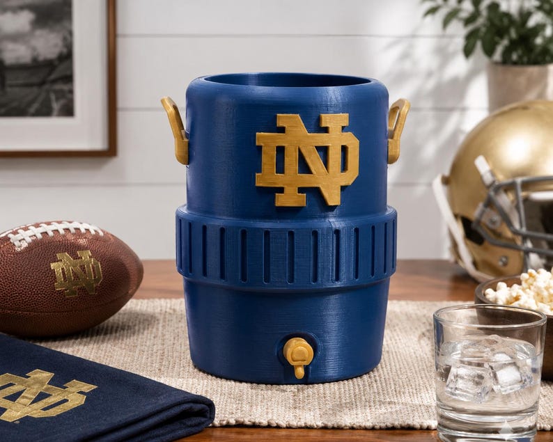 May include: A blue cylindrical beverage cooler featuring a gold Notre Dame ND logo on the front, two gold side handles, and a gold spigot at the base. The cooler has a ribbed texture around the middle section and a smooth finish on the top and bottom.