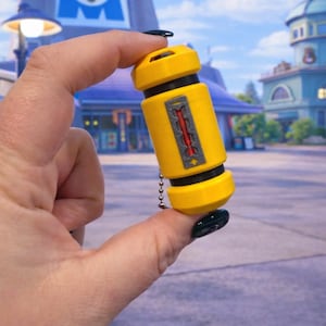 May include: A yellow cylindrical keychain with black accents and a decorative panel featuring a red design. The keychain is held up against a backdrop of a building with a large blue "M" logo.