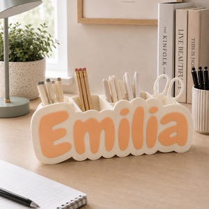 May include: A cream-coloured desk organiser with a scalloped edge featuring the name Emilia in raised, peach-coloured letters. The organiser has multiple compartments holding various pens, pencils, and a pair of white scissors.