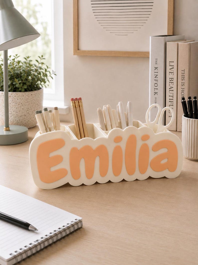 May include: A cream-coloured desk organiser with a scalloped edge featuring the name Emilia in raised, peach-coloured letters. The organiser has multiple compartments holding various pens, pencils, and a pair of white scissors.