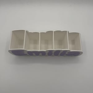 May include: A white desk organiser with five rectangular compartments. The front panel features the name Emilia in raised, light purple, rounded lettering. The structure is made of a solid material with a clean, smooth finish and open top.
