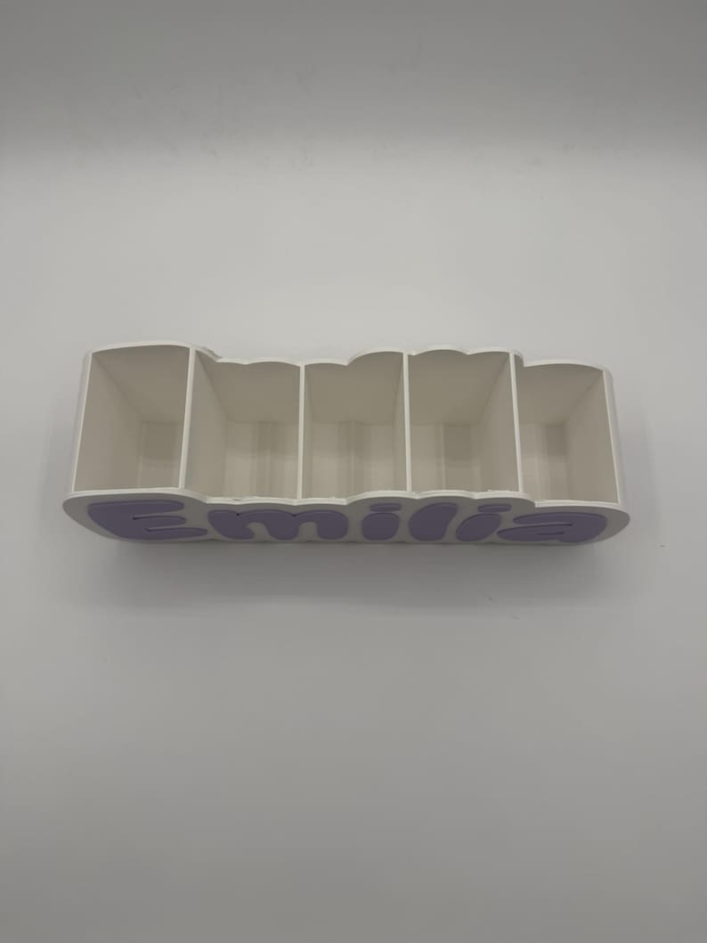 May include: A white desk organiser with five rectangular compartments. The front panel features the name Emilia in raised, light purple, rounded lettering. The structure is made of a solid material with a clean, smooth finish and open top.