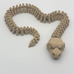 May include: A tan-colored, articulated snake toy with a detailed skeletal design. The snake's head features a stylized, cartoonish face with closed eyes and a slightly menacing expression. The segmented body is curved.