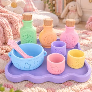 May include: A set of pastel-colored silicone play items on a purple wavy tray, including a light blue bowl with a pink spoon, three bottles with cork-style stoppers, and three small cups, all featuring embossed celestial and floral motifs.