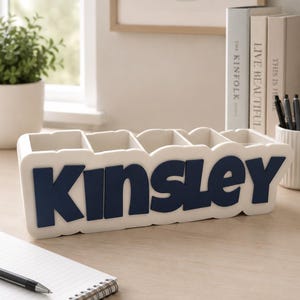 May include: A white rectangular desk organiser with five compartments, featuring the name Kinsley in large, bold, dark blue raised lettering on the front. The organiser has a smooth, matte finish and a clean, structured design for holding pens.