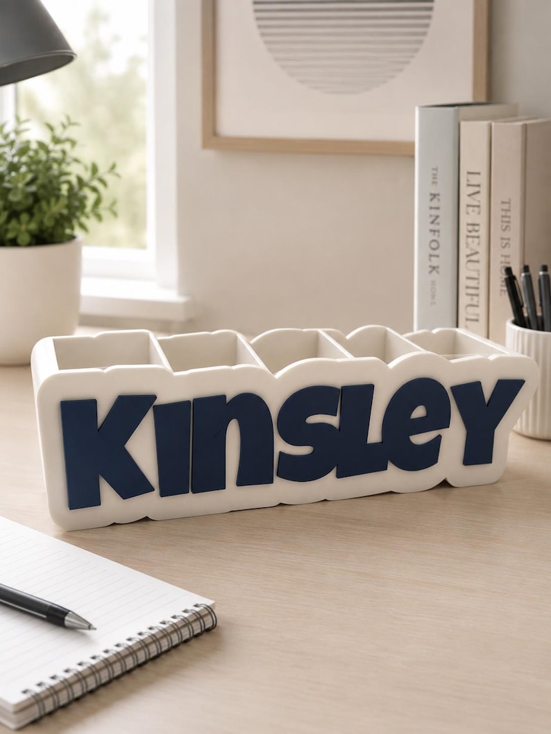 May include: A white rectangular desk organiser with five compartments, featuring the name Kinsley in large, bold, dark blue raised lettering on the front. The organiser has a smooth, matte finish and a clean, structured design for holding pens.