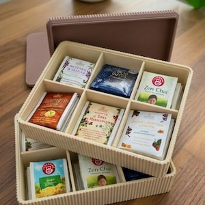 May include: A beige tea organizer with multiple compartments, filled with various tea bags. The tea bags feature colorful packaging with text such as "Rooibos" and "Zen Chai". A matching lid and a white mug are also visible.
