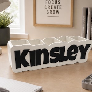 May include: A white rectangular desk organiser with five vertical compartments. The front of the organiser features the name KINSLEY in large, bold, black capital letters. The structure is made of a smooth material with a clean, modern finish.
