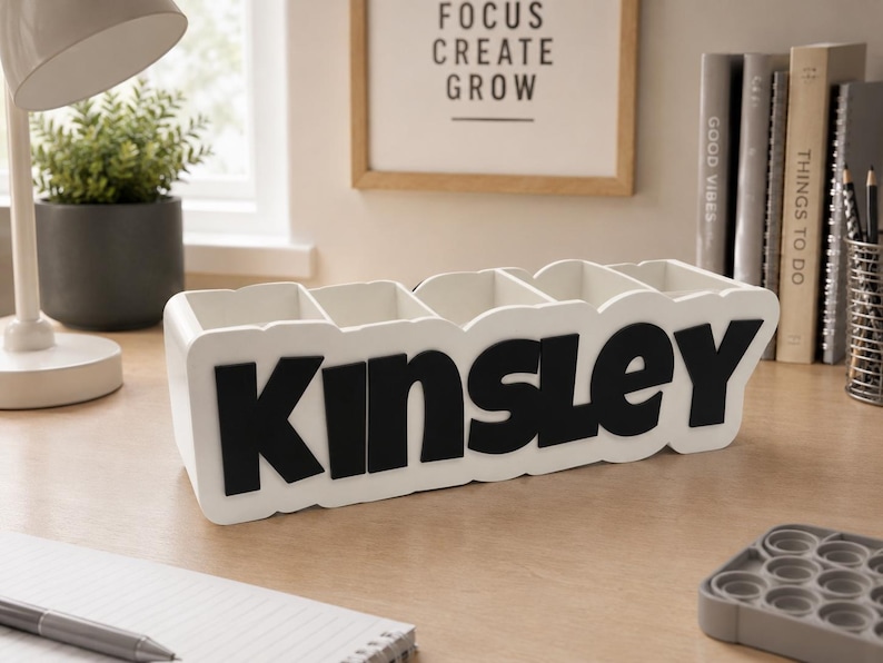 May include: A white rectangular desk organiser with five vertical compartments. The front of the organiser features the name KINSLEY in large, bold, black capital letters. The structure is made of a smooth material with a clean, modern finish.