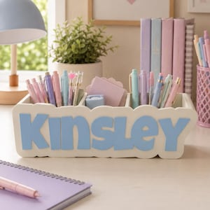 May include: A white rectangular desk organiser with scalloped edges featuring the name Kinsley in large, light blue, raised letters on the front. The organiser has three compartments filled with various pastel-coloured pens, pencils, and a stack of light pink sticky notes.