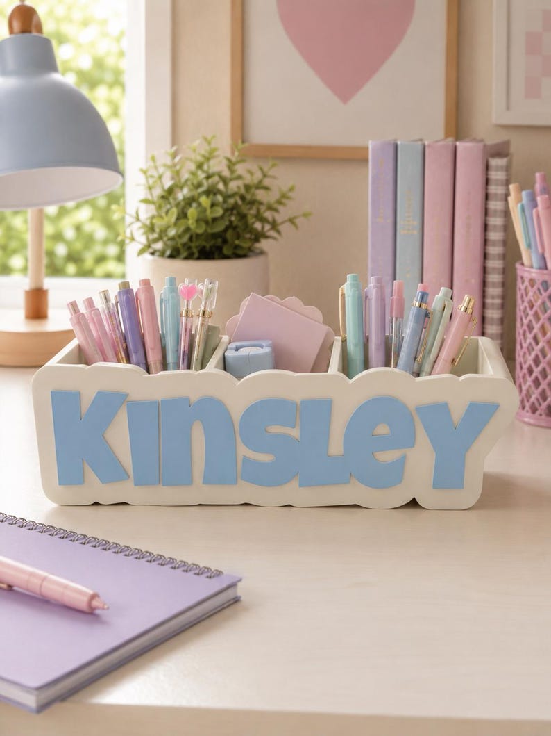 May include: A white rectangular desk organiser with scalloped edges featuring the name Kinsley in large, light blue, raised letters on the front. The organiser has three compartments filled with various pastel-coloured pens, pencils, and a stack of light pink sticky notes.