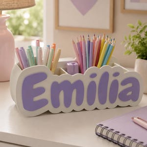 May include: A white desk organiser with multiple compartments for stationery, featuring the name Emilia in large, raised purple letters on the front. The organiser is filled with an assortment of colourful pencils and pens in pastel shades.