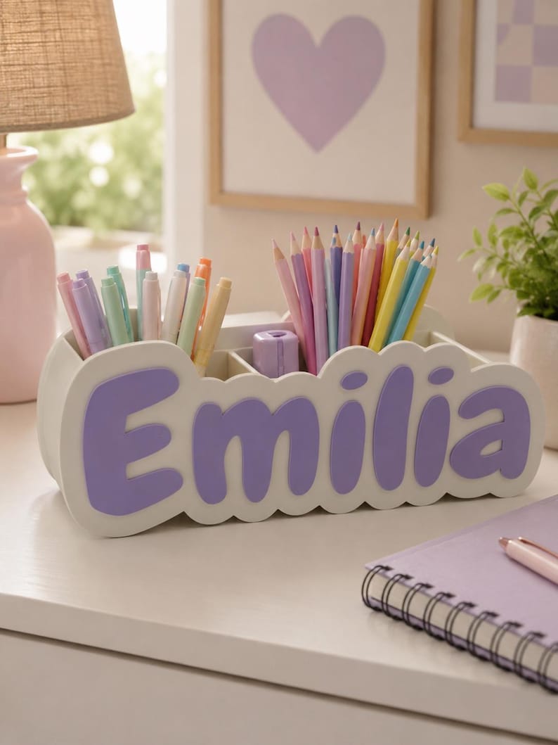 May include: A white desk organiser with multiple compartments for stationery, featuring the name Emilia in large, raised purple letters on the front. The organiser is filled with an assortment of colourful pencils and pens in pastel shades.