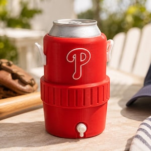 Może przedstawiać: Czerwony chłodziarka do napoj&oacute;w zaprojektowana na standardową aluminiową puszkę, z białym logo Philadelphia Phillies z przodu, białym kranikiem u podstawy i dwoma małymi białymi uchwytami bocznymi, z widoczną metaliczną srebrną g&oacute;rą puszki w otworze.