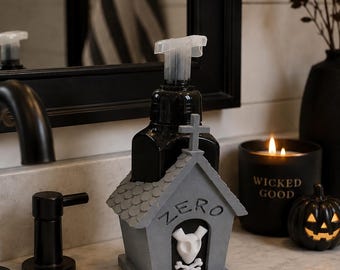May include: A black soap dispenser pump sits inside a gray, house-shaped holder with a shingled roof and a cross on top. The front of the holder features the word ZERO in raised letters above a white ghost-like figure with crossed bones.