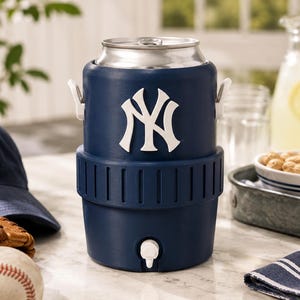 Może przedstawiać: Granatowa chłodziarka do napoj&oacute;w w kształcie piłki baseballowej, z białym logo New York Yankees z przodu, srebrną metalową pokrywą, białym kranikiem u podstawy oraz teksturowanym środkowym pasem z dwoma białymi uchwytami po bokach do przenoszenia.