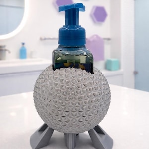 May include: A gray, geometric sphere-shaped soap dispenser holder with a blue pump. The holder has a textured surface and is supported by three gray legs. The dispenser holds a clear bottle with green liquid.