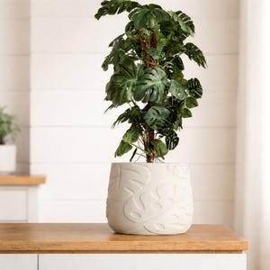 May include: A tall, green artificial plant with large, split leaves in a white, textured pot. The pot features a repeating leaf pattern. The plant is placed on a wooden countertop against a white background.