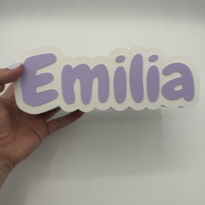 May include: A custom name sign featuring the name Emilia in bold, rounded, light purple letters. The letters are layered on top of a white, scalloped-edge backing that follows the contour of the text, creating a dimensional effect.