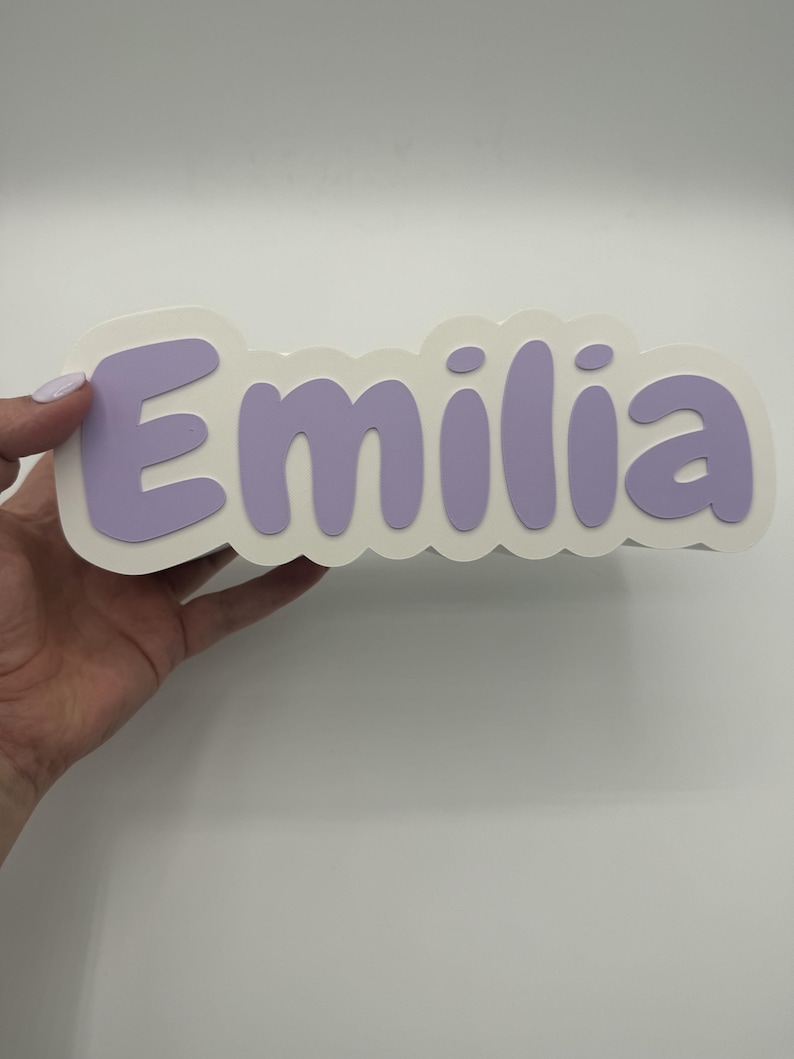 May include: A custom name sign featuring the name Emilia in bold, rounded, light purple letters. The letters are layered on top of a white, scalloped-edge backing that follows the contour of the text, creating a dimensional effect.