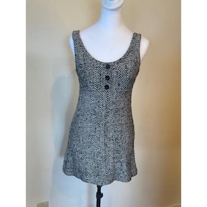 May include: A sleeveless mini dress featuring a black and white herringbone pattern. The dress has a scoop neckline and a vertical row of three black buttons centered on the upper bodice, creating a structured look with a fitted waistline.