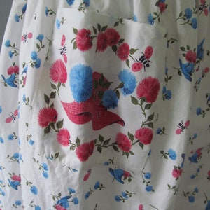 May include: White cotton apron with a large pocket featuring a floral print of pink and blue flowers, green leaves, and small red ladybugs. The pocket has a red and white hat design in the center.