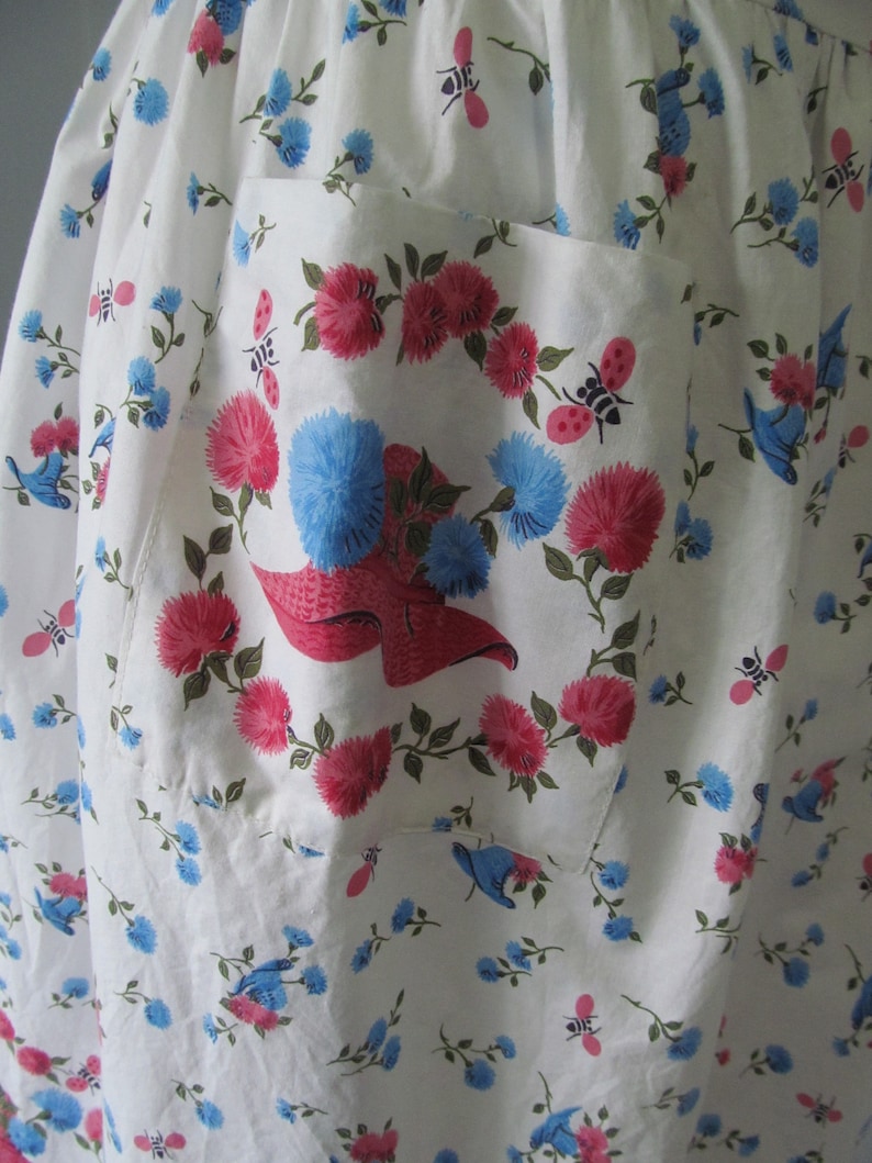 May include: White cotton apron with a large pocket featuring a floral print of pink and blue flowers, green leaves, and small red ladybugs. The pocket has a red and white hat design in the center.