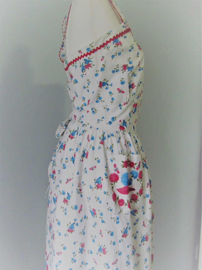 May include: A white dress with a blue and pink floral print. The dress has a halter neckline and a red trim around the neckline. The dress has a single pocket on the right side.