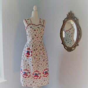 May include: A white sleeveless dress with a red and blue floral print. The dress has a square neckline and a fitted bodice. The skirt is full and flows to the knees. The dress is on a white mannequin in a room with a light blue wall.