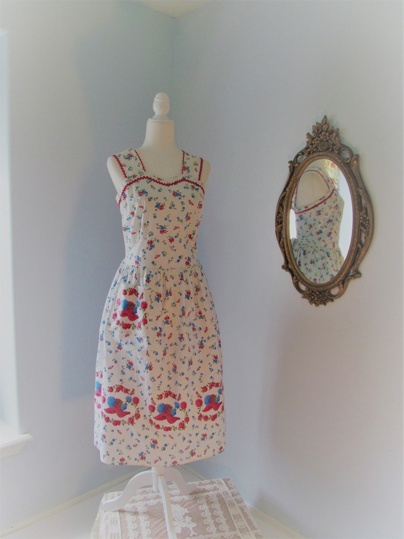 May include: A white sleeveless dress with a red and blue floral print. The dress has a square neckline and a fitted bodice. The skirt is full and flows to the knees. The dress is on a white mannequin in a room with a light blue wall.