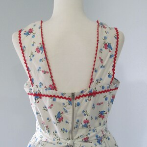 May include: White sleeveless dress with a floral print featuring blue and pink flowers. The dress has a red scalloped trim around the neckline and armholes.