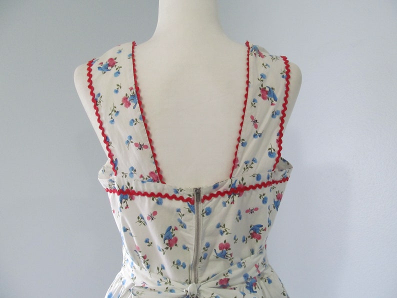 May include: White sleeveless dress with a floral print featuring blue and pink flowers. The dress has a red scalloped trim around the neckline and armholes.