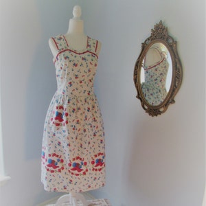 May include: A white sleeveless dress with a red and blue floral print. The dress has a scalloped neckline and a fitted bodice. The dress is on a white mannequin and is reflected in a gold oval mirror.