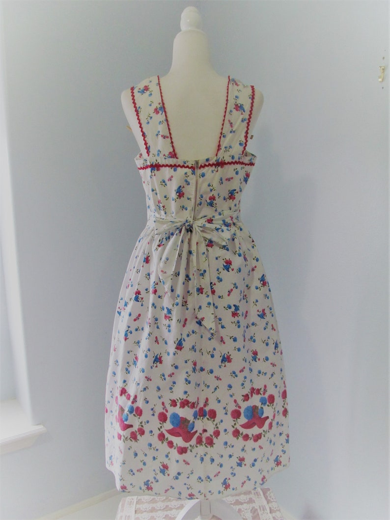 May include: A white sleeveless dress with a floral print featuring red, blue, and white flowers. The dress has a red trim around the neckline and a bow at the back.