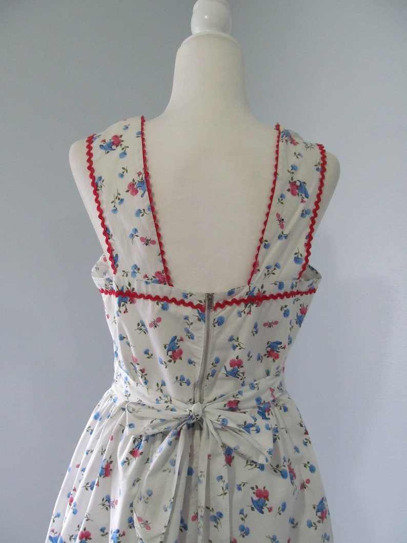 May include: A white dress with a blue and pink floral print. The dress has a red trim around the neckline and a bow at the back.