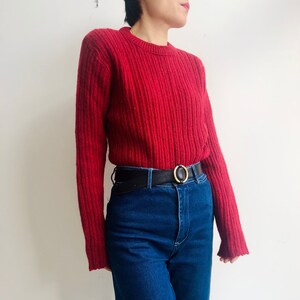 May include: A red ribbed knit sweater with long sleeves and a crew neck. The sweater is paired with high-waisted, dark blue denim jeans and a black belt with a gold-colored buckle. The outfit is simple and classic.