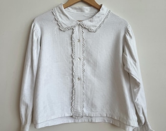 May include: White long-sleeved blouse with a Peter Pan collar and delicate lace trim. The blouse features small, round buttons down the front and lace detailing on the cuffs. The garment is hanging on a wooden hanger.