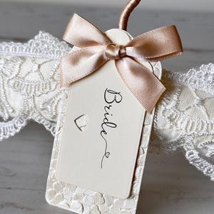 Op de afbeelding: Een rechthoekig, cr&egrave;mekleurig label met het woord Bride in zwarte schrijfletters, voorzien van een kleine hartvormige uitsparing en een champagnekleurige satijnen strik. Het label ligt op een witte kanten band met hartjes.