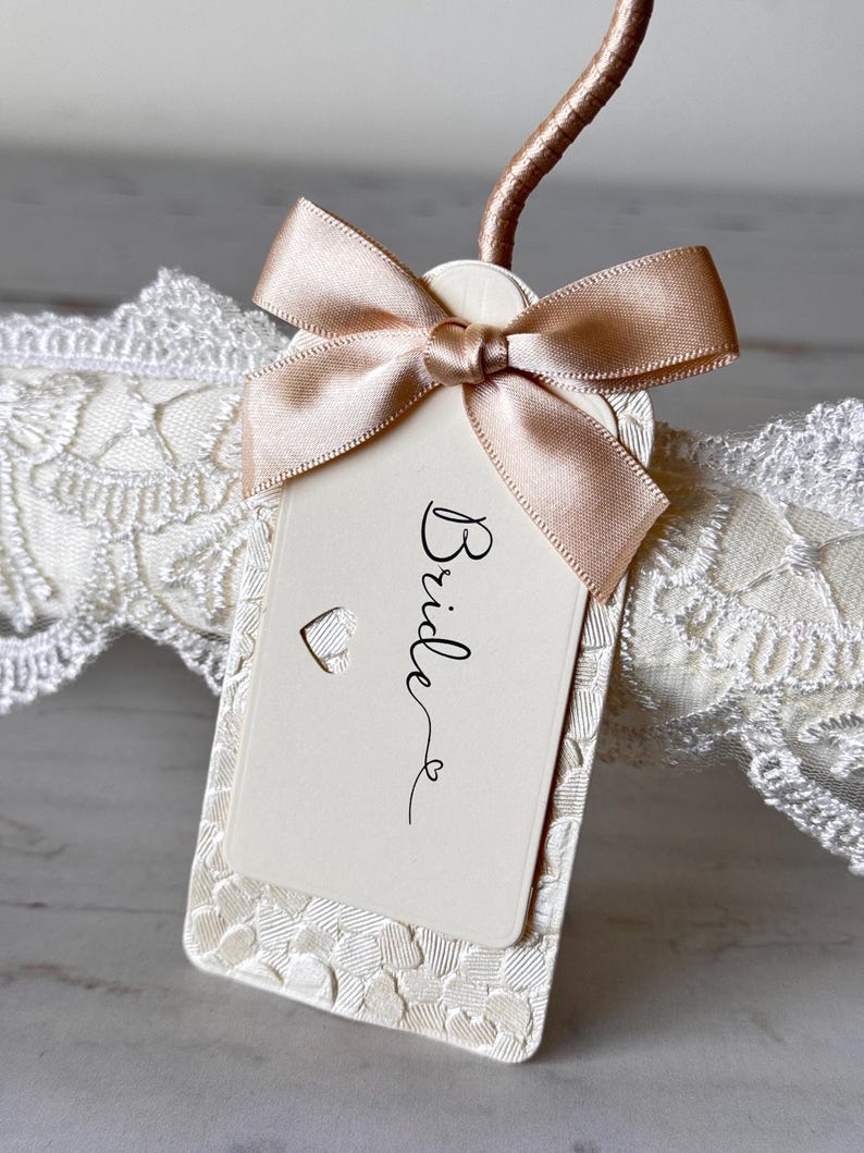 Op de afbeelding: Een rechthoekig, cr&egrave;mekleurig label met het woord Bride in zwarte schrijfletters, voorzien van een kleine hartvormige uitsparing en een champagnekleurige satijnen strik. Het label ligt op een witte kanten band met hartjes.
