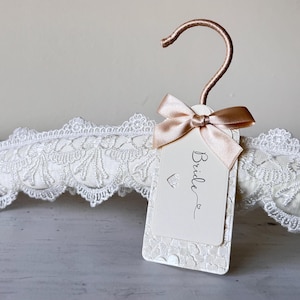 Op de afbeelding: Een gewatteerde kledinghanger bekleed met witte stof en een decoratieve witte kanten rand. De hanger heeft een ros&eacute;gouden metalen haak en een rechthoekig gebroken wit label vastgemaakt met een satijnen lint. Het woord Bride is in het zwart gedrukt.