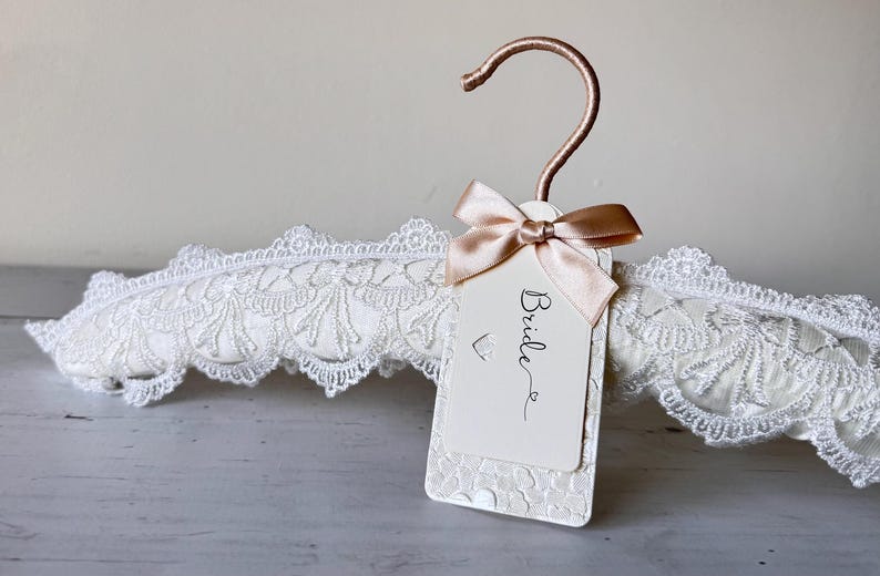 Op de afbeelding: Een gewatteerde kledinghanger bekleed met witte stof en een decoratieve witte kanten rand. De hanger heeft een ros&eacute;gouden metalen haak en een rechthoekig gebroken wit label vastgemaakt met een satijnen lint. Het woord Bride is in het zwart gedrukt.