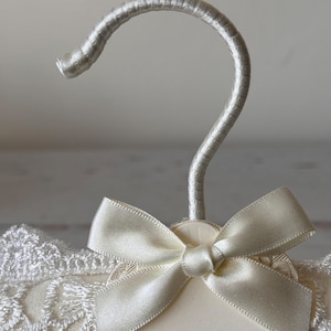 Op de afbeelding: Een gevoerde kledinghanger bekleed met cr&egrave;mekleurige stof met een witte kanten overlay. De hanger heeft een zilverkleurige haak omwikkeld met bijpassende stof en een cr&egrave;mekleurige satijnen strik aan de basis van de haak, met een label Bride.